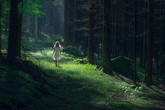 Beautiful young woman wearing elegant white dress standing in the forest with rays of sunlight beaming through the leaves of the trees