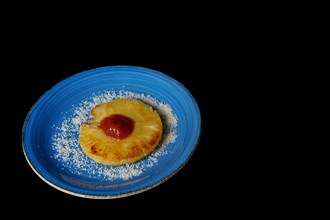 Close-up of a grilled natural pineapple with grated coconut on a blue plate on a black background