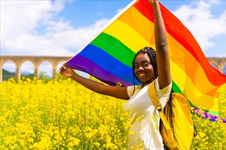 A smiling black ethnic girl with braids holding the LGBT flag in a field of yellow flowers