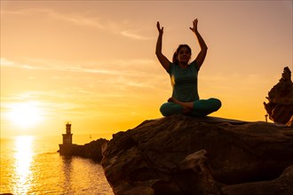 A woman doing meditation and yoga exercises at sunset next to a lighthouse in the sea