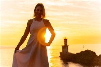Portrait of a blonde woman smiling in a white dress at sunset next to a lighthouse in the sea