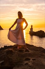 Portrait of a blonde woman in a white dress at sunset next to a lighthouse in the sea