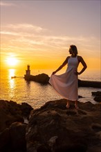 A blonde woman in a white dress at sunset next to a lighthouse in the sea