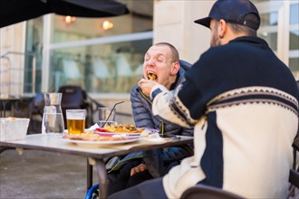 A disabled person eating on the terrace of a restaurant and a friend helping him eat