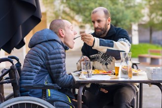 A disabled person eating on the terrace of a restaurant and a friend helping him eat