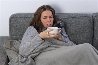 Sick woman covered with a blanket drinking an infusion on the couch in her home