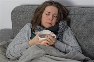 Sick woman asleep on couch covered with a blanket with a cup of tea in her hand