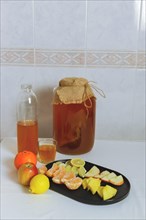 Large glass bottle with fermented kombucha tea on the white kitchen table and assorted fruit for taste