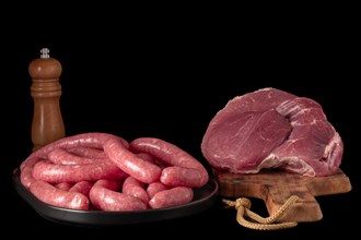 Fresh raw sausages on a black tray and Veal sirloin steak raw on a wooden board isolated on a black background