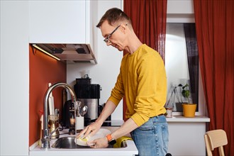 Middle-aged man in spectacles washing dishes in kitchen in the evening
