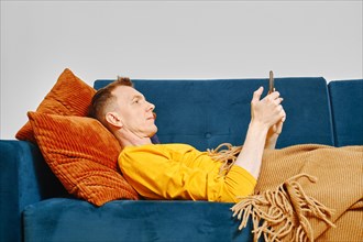 Man rests on couch covered with blanket and reads news on smartphone
