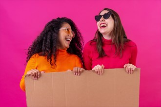 Two smiling multiethnic female friends holding a cardboard sign over a pink background