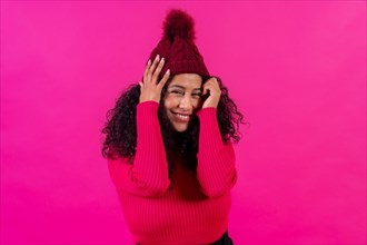 Curly-haired woman in a woolen cap on a pink background smiling shyly