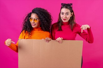 Two female friends smiling and pointing at the cardboard sign on a pink background