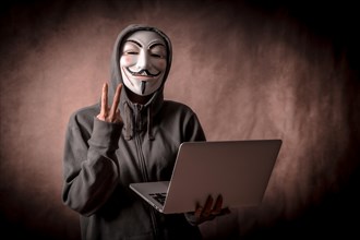 Hacker with anonymous mask with a laptop making the victory symbol