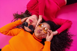 Two multi-ethnic women in sunglasses lying down smiling on a pink background