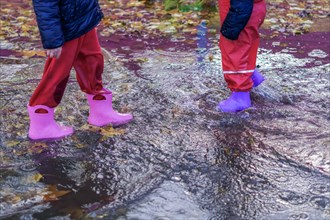 Two children walking through a puddle of water with their boots on a rainy day