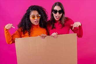 Two female friends smiling and pointing at the cardboard sign on a pink background