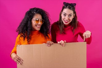 Two multiethnic female friends smiling and pointing at cardboard sign over pink background