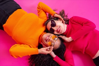 Two multi-ethnic women in sunglasses lying down laughing on a pink background