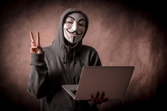 Hacker with anonymous mask with a laptop making the victory symbol