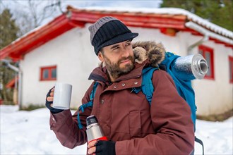 Man drinking coffee from a hot thermos in winter in the snow next to a cabin after sleeping in it
