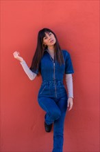 Gorgeous young brunette girl smiling leaning against a wall in a blue denim outfit