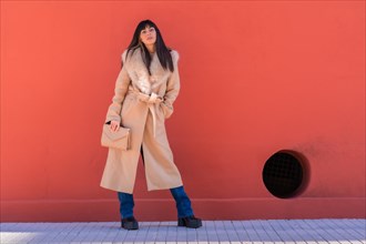 Posing of a smiling brunette model in a winter jacket leaning against a wall with a bag