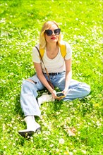 Portrait of a blonde tourist woman in a hat and sunglasses sitting on the grass in spring next to daisies