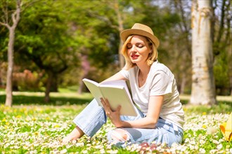 A young blonde girl in a hat reading a book in spring in a park in the city