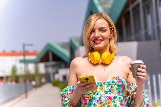 Portrait of a blonde woman walking with the phone through the city with yellow headphones and a take away coffee