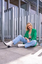 Portrait of a blonde model woman in a green jacket sitting on the floor in a building in the city