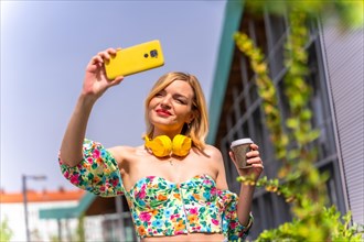 A blonde woman taking a selfie with her phone in the city with yellow headphones and a take away coffee