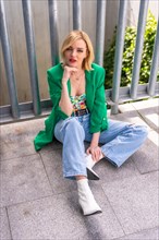 Photo from above of a blonde model woman in a green jacket sitting on the floor in a building in the city
