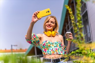 Portrait of a blonde woman taking a selfie with her phone in the city with yellow headphones and a take away coffee