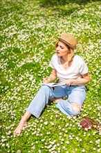 A young blonde girl in a hat writing poems in a notebook in spring in a park in the city