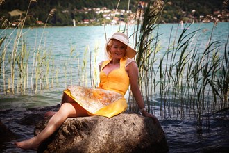 Elegant woman in dirndl and sun hat sits smiling on a rock in a lake