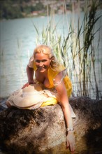 Elegant woman with dirndl sits smiling on a stone in the lake and holds one hand in the water
