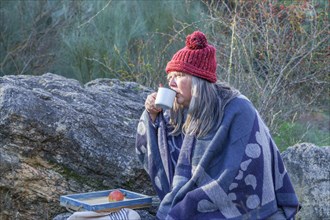 Woman with white hair and red cap drinking coffee in a field with a tray of fruit