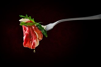 Piece of marbled beef carpaccio on fork with dripping olive oil over black background