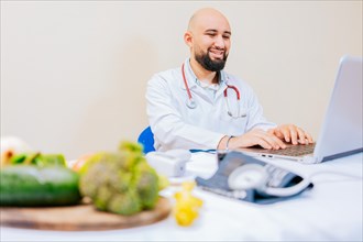 Smiling nutritionist with laptop at desk. Nutritionist doctor using laptop at workplace. Bearded nutritionist doctor working on laptop at desk