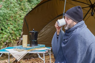 Bearded man covered with a camping blanket sipping a cup of coffee