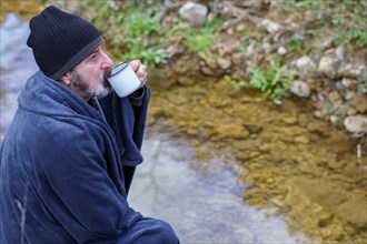 Bearded man covered with a blanket drinking coffee on the river bank