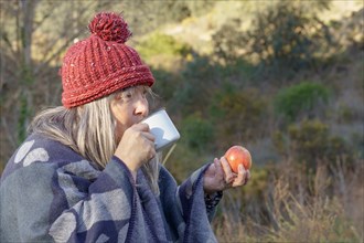 Woman with white hair and red cap drinking coffee and eating an apple in a field with trees in the background