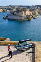 View over former parade ground next to Upper Barraka Gardens in La Valletta with salute cannon next to two soldiers in historical uniform on historical Fort St. Angelo on Birgu Peninsula