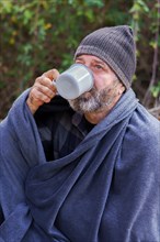 Bearded man covered with a camping blanket sipping a cup of coffee