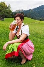 Young Bavarian woman in a colourful embroidered traditional dirndl kneels on the lawn in a lush green alpine pasture drinking from a large mug of beer as she celebrates Oktoberfest