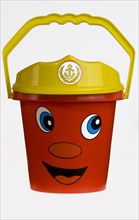 A bright red plastic childs bucket with a yellow handle and a smiley face