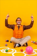 Juggler in waistcoat and with painted face sitting with the juggling objects on a yellow background