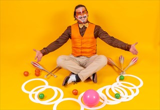 Portrait of a juggler in a vest and makeup sitting with the juggling objects on a yellow background